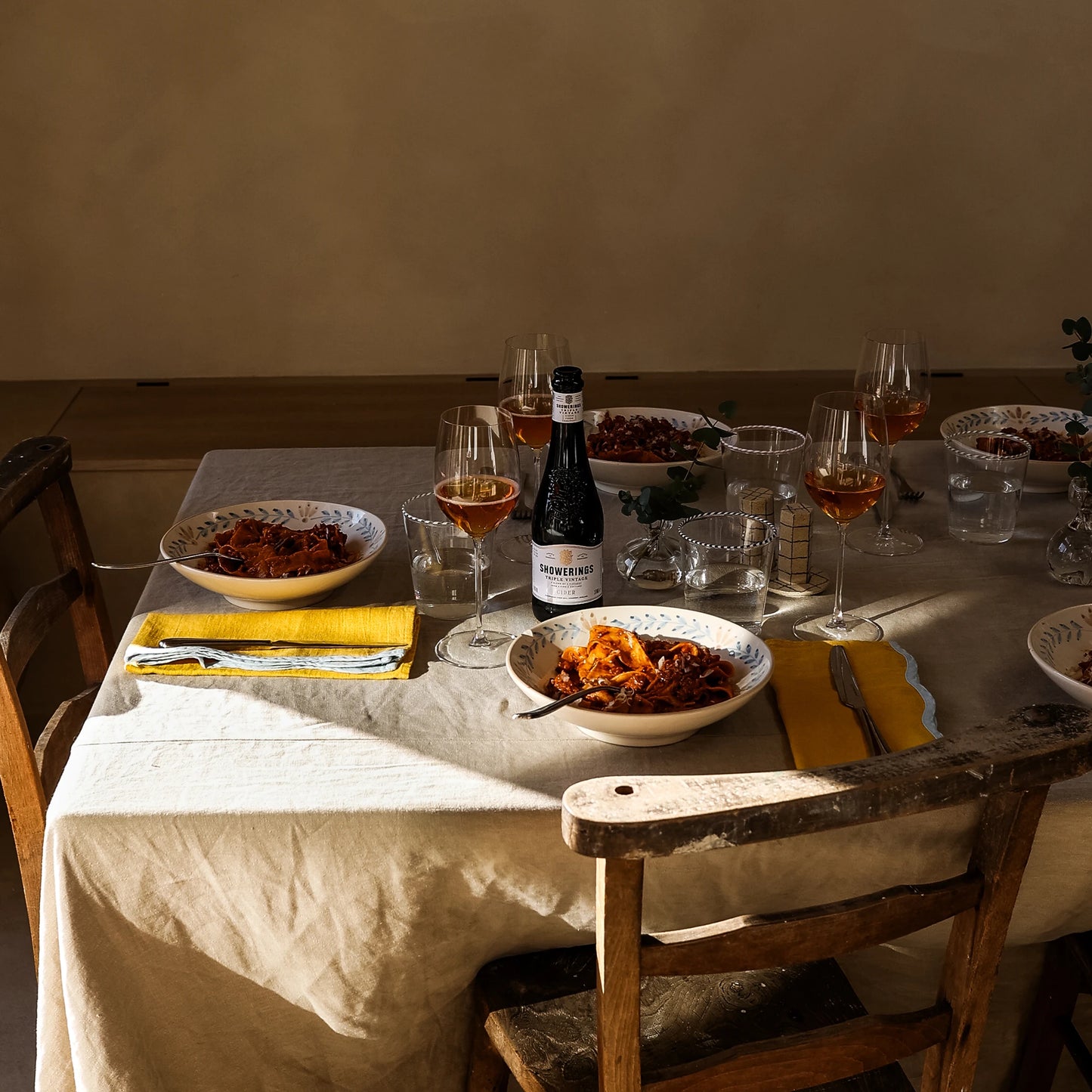 
                  
                    Dining table covered by tablecloth, with bowls of food, cutlery, wine glasses, cider and sunlight  
                  
                